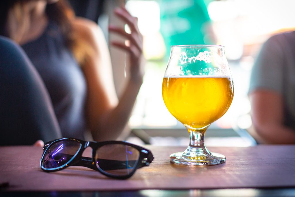 Ein halbvolles Bierglas steht auf einem Tisch, daneben liegt eine Brille.