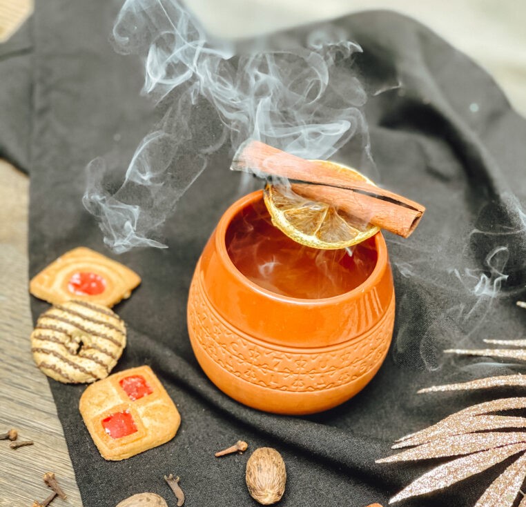 Eine Orangene Tasse, die eine heißes, dampfendes Getränk enthält. Quer über der Tasse liegt eine Zimtstange.