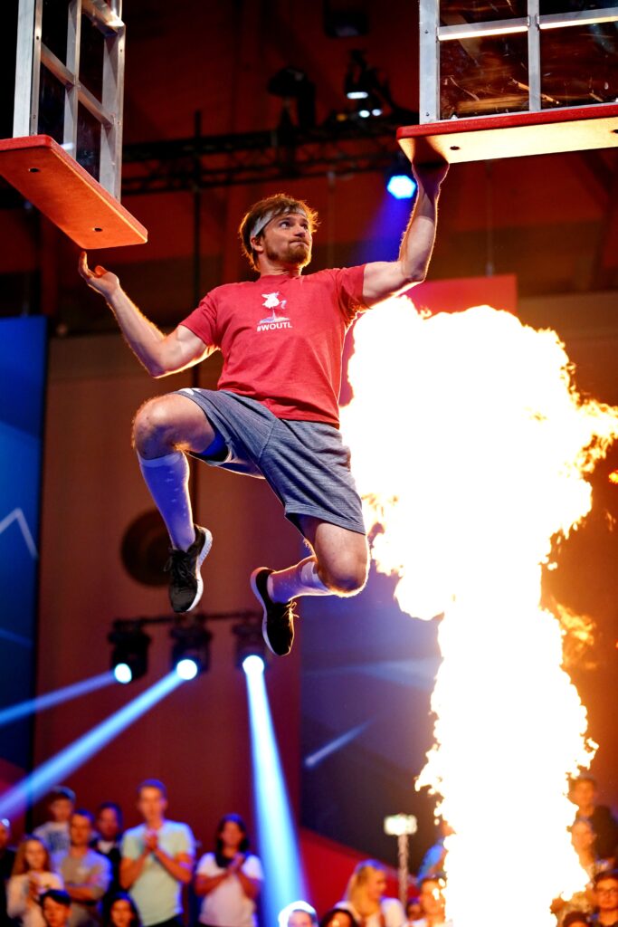 Mann bei der Absolvierung eines Ninja Warrior Parkours. Hinter ihm geht eine Flamme auf