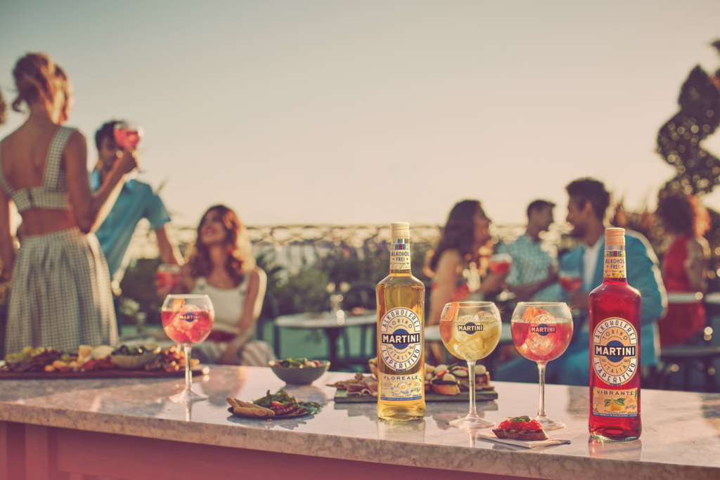 Menschen sitzen beisammen und trinken amüsiert einen alkoholfreien Martini, den es in zwei Geschmackssorten gibt. Im Vordergrund stehen die zwei Martini-Flaschen auf einer Steinplatte.