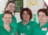 Frau mittleren Alters mit roten Haaren und grüner Uniform. Sie ist umgeben von ihren Mitarbeiterinnen.