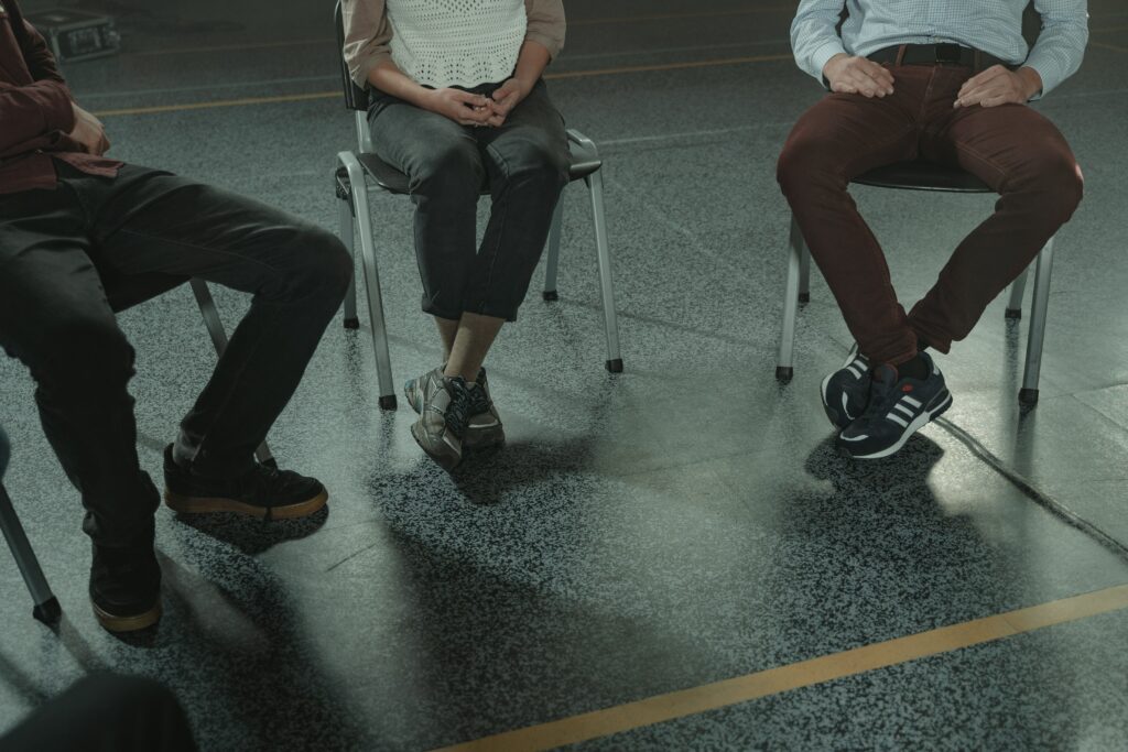 Das Bild zeigt drei Personen, die jeweils auf einem Stuhl sitzen. Ihre Gesichter sind nicht zu sehen.