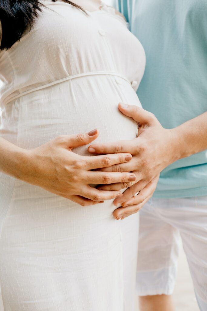 Auf dem Bild ist eine schwangere Frau zu sehen. Ihr Partner und sie legen die Hände auf dem Bauch.