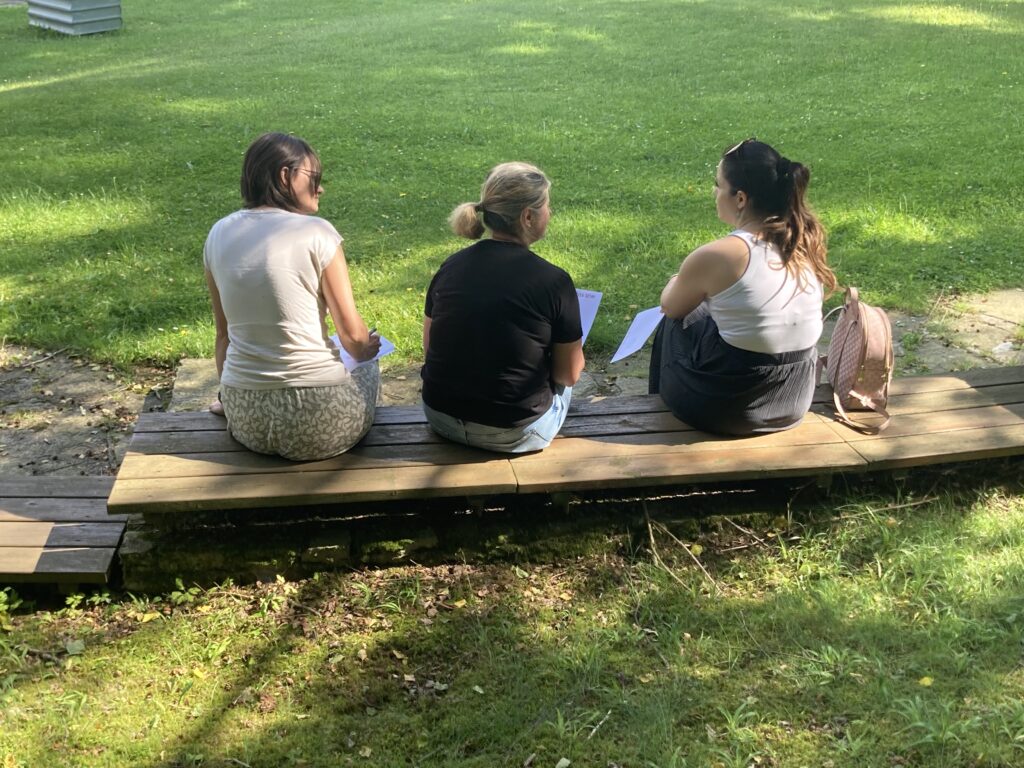 Drei Frauen jung bis mittleren Alters, sitzen auf einer Bank auf einer Wiese.